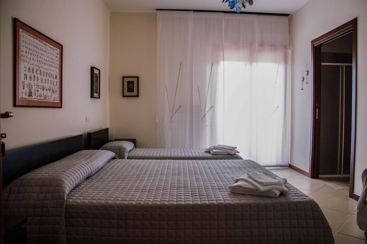 A hotel room with two beds, a large window with sheer curtains, a framed picture on the wall, and a door to the right.