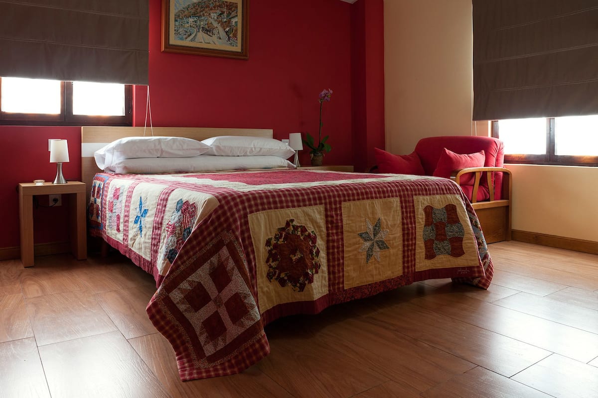 A bedroom with a large bed, a nightstand with a lamp, a chair, and a window with blinds.