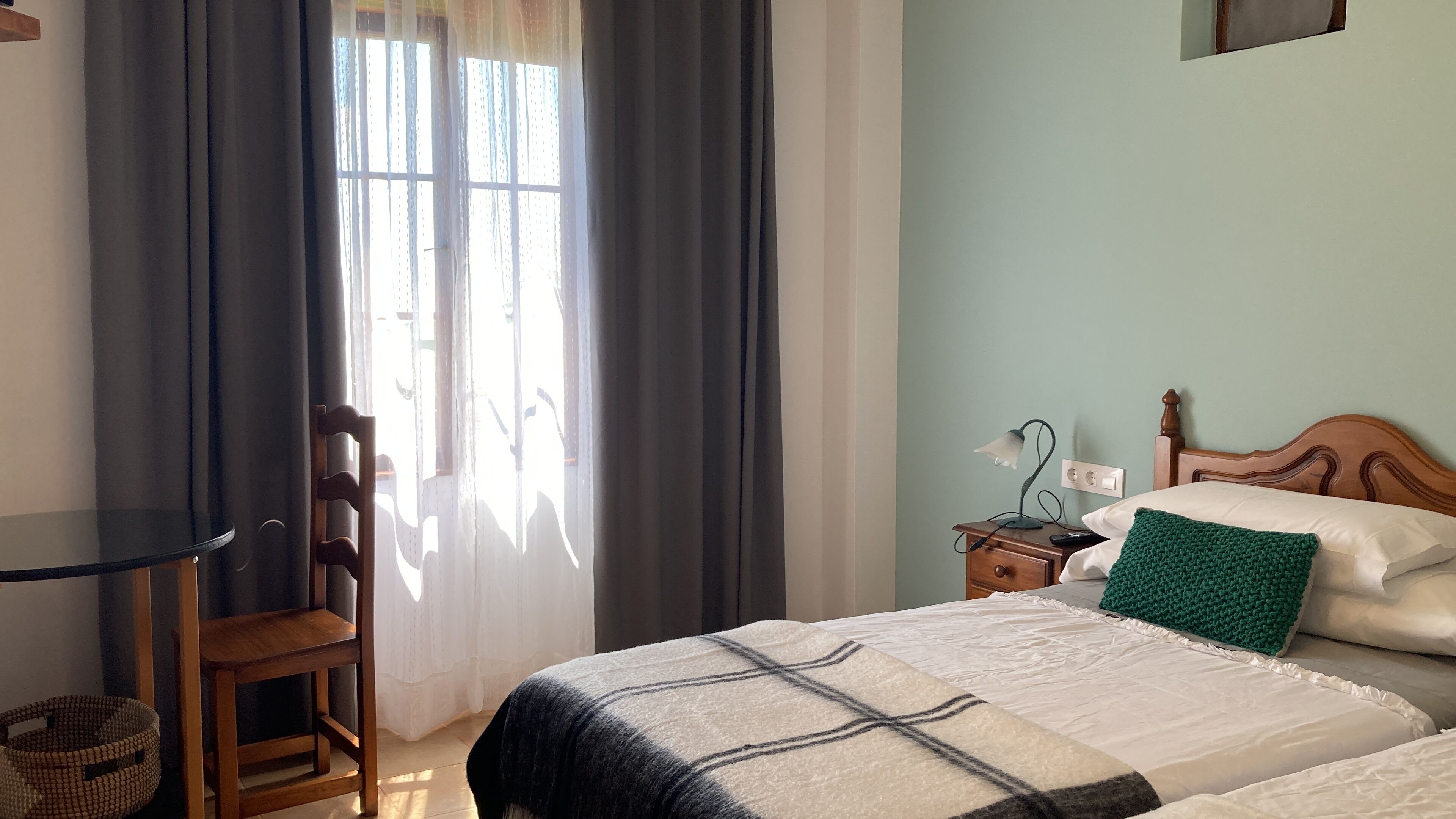 Un dormitorio con cama, una silla, una mesita de noche con lámpara y una ventana con cortinas.