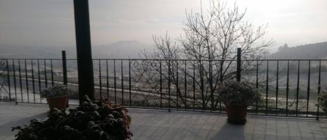 Un balcón con barandilla de metal, plantas en macetas y vistas a un paisaje con árboles y colinas al lejano.