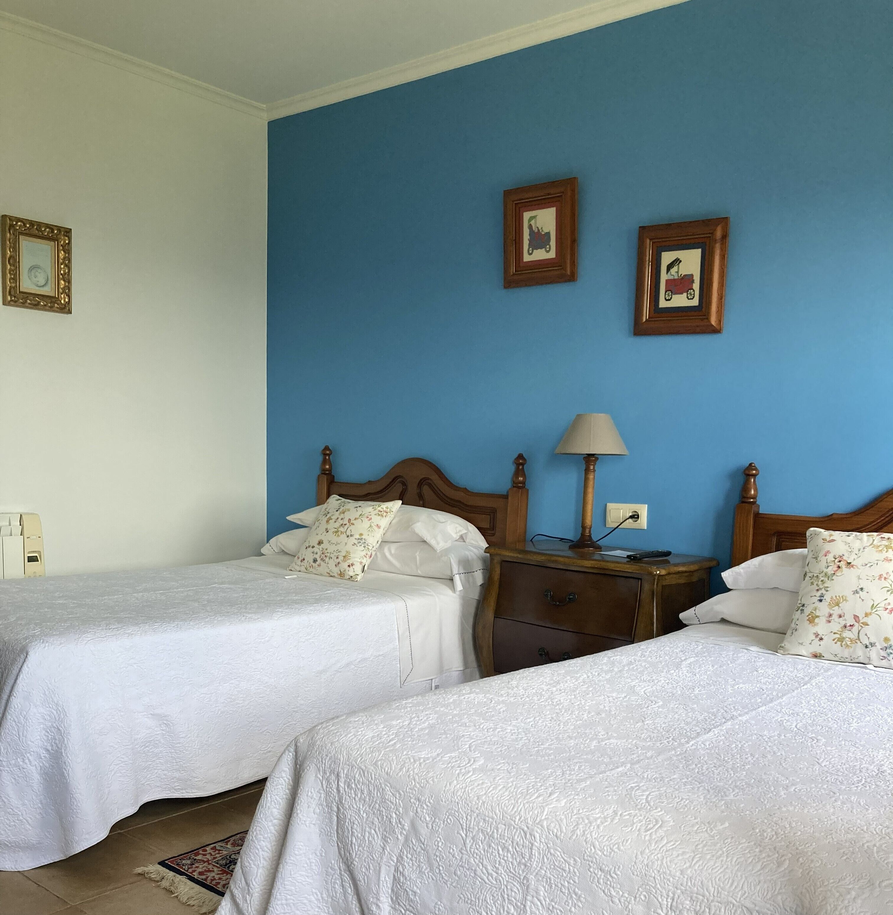 Habitación de hotel con dos camas, una mesita de noche con lámpara y dos cuadros enmarcados en la pared.