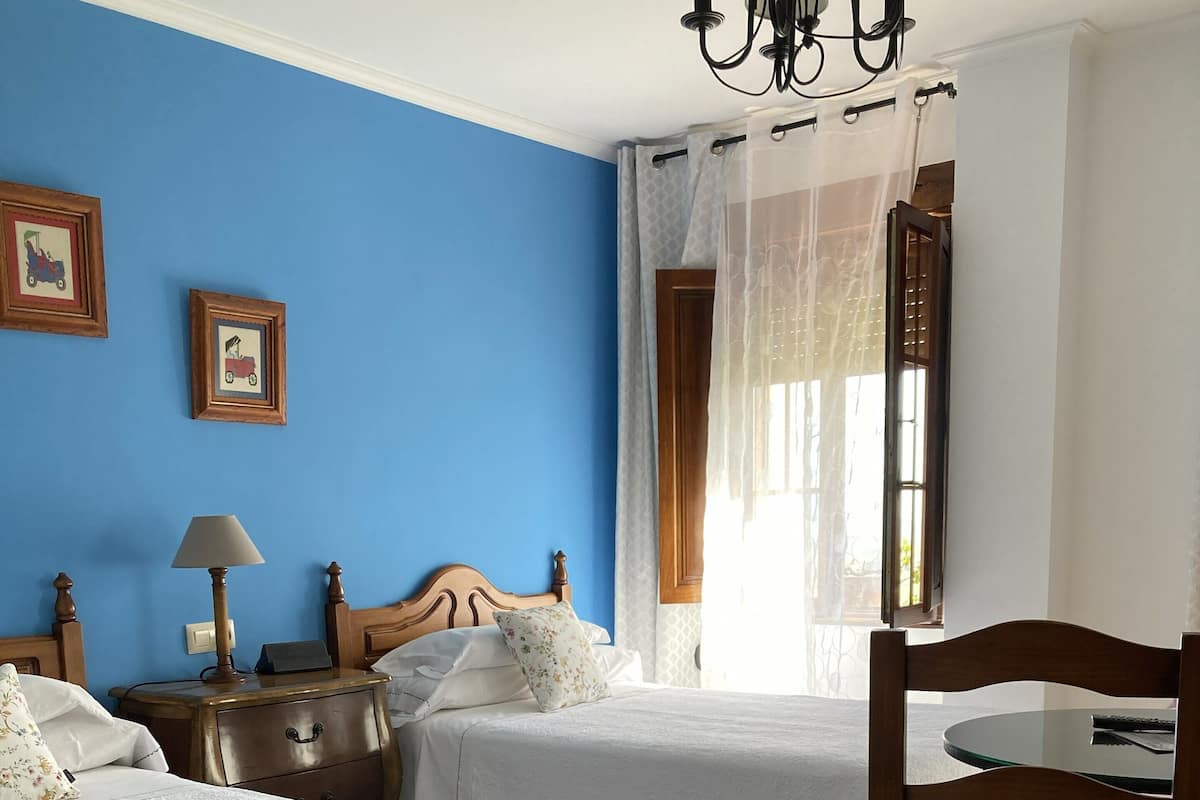 A bedroom with two beds, a nightstand, a lamp, a chair, a window with curtains, and a chandelier.