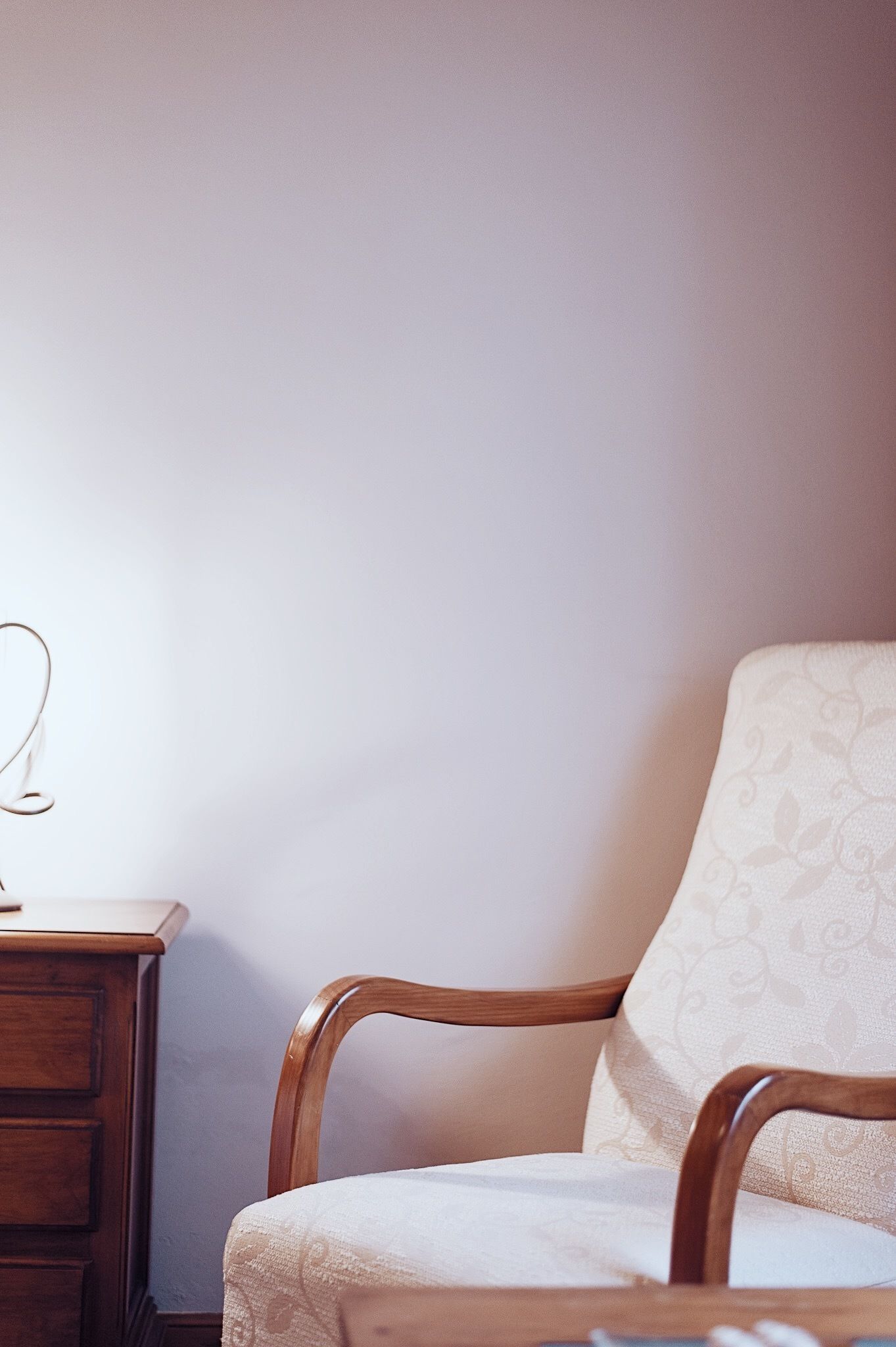 Une chaise en bois avec un coussin à motifs, placée devant un mur uni, une table de chevet en bois avec une lampe, et une commode en bois dont on ne voit qu’une partie.