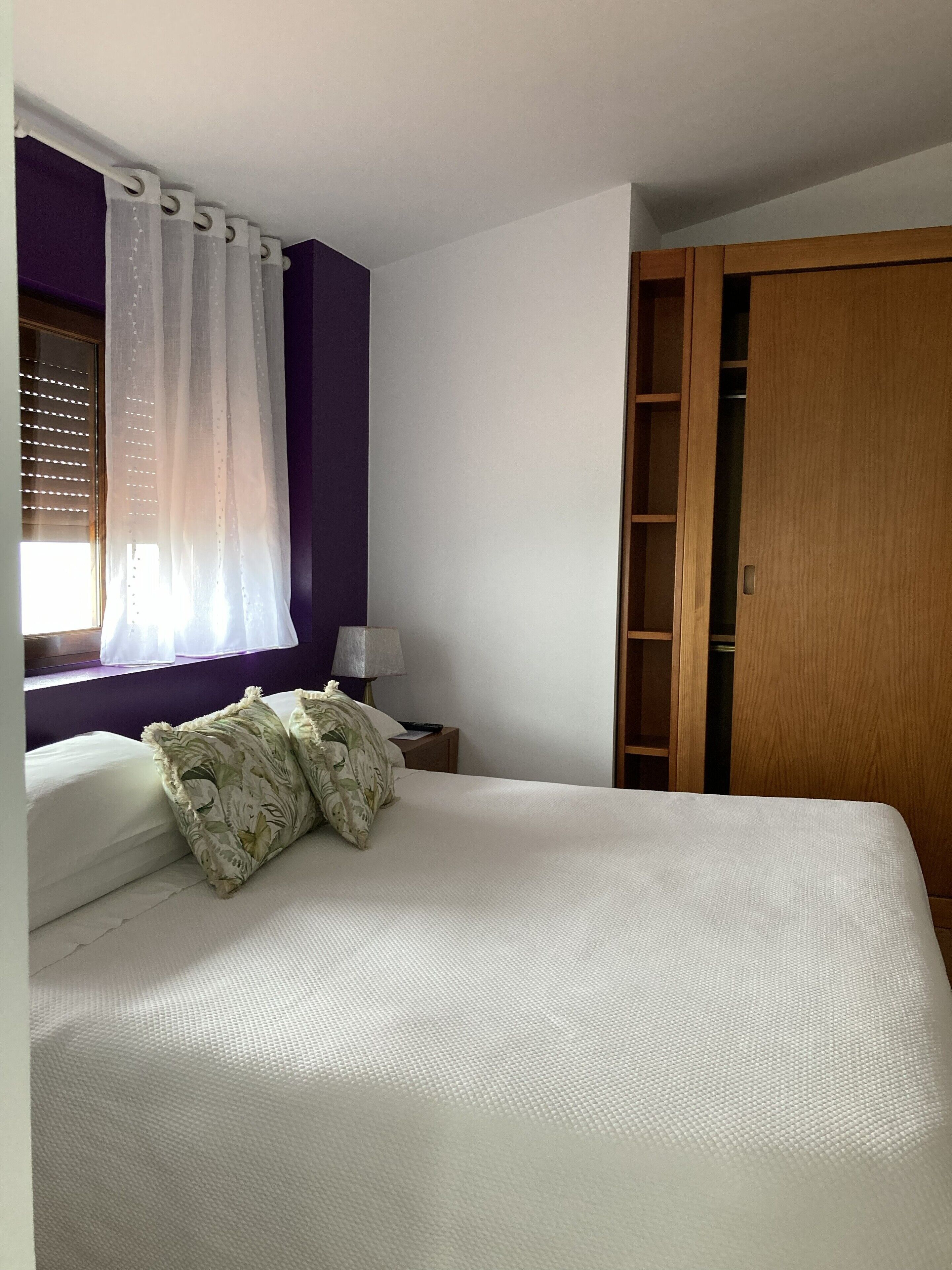 A bedroom with a bed, a window with blinds, a nightstand with a lamp, and a wooden wardrobe.