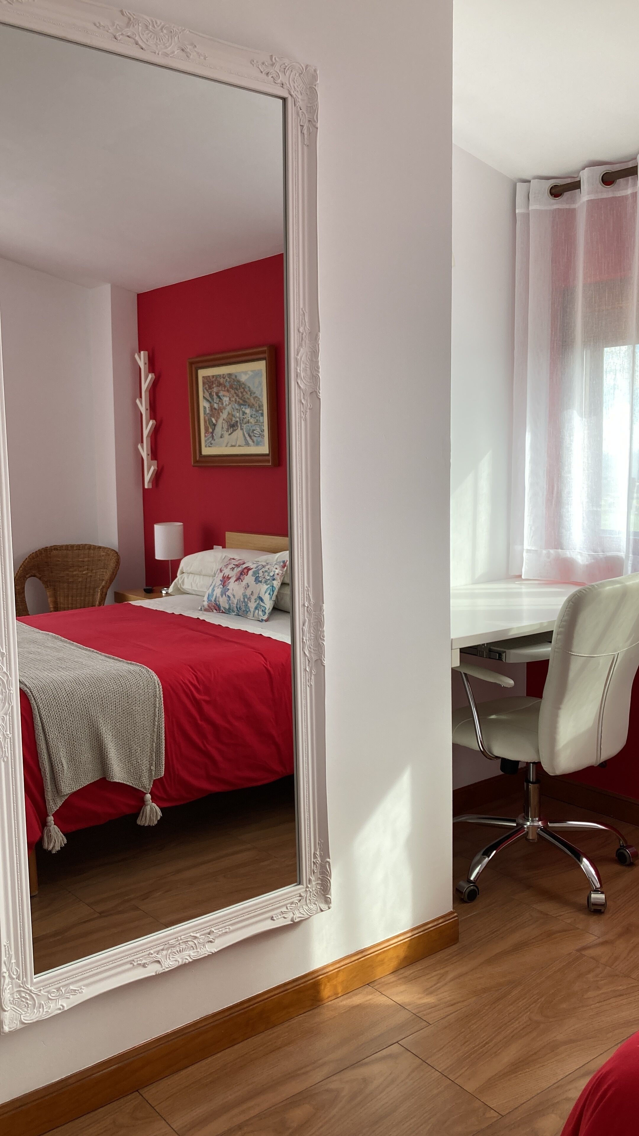 Una habitación con un espejo grande, una cama con ropa de cama roja, un escritorio con una silla, una ventana con cortinas traslúcidas y un cuadro en la pared.