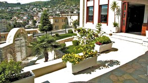 A balcony with a view of a cityscape, including a hill with buildings and a water tower. There are potted plants and a palm tree on the balcony. - Sharm Hotel (Gjirokastër)