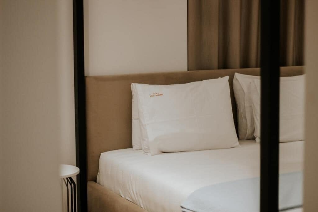 A neatly made bed with white linens and pillows, a bedside table with a lamp, and a window with curtains.