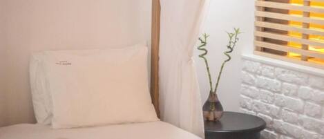 A bedroom with a bed, a bedside table, a vase with a plant, and a window with blinds.