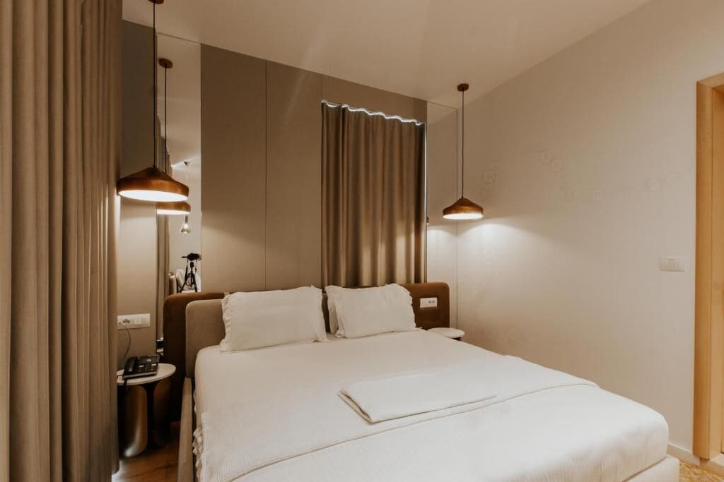 Una habitación de hotel moderna con una cama grande, dos lámparas colgantes, una mesita de noche con lámpara y una cortina.