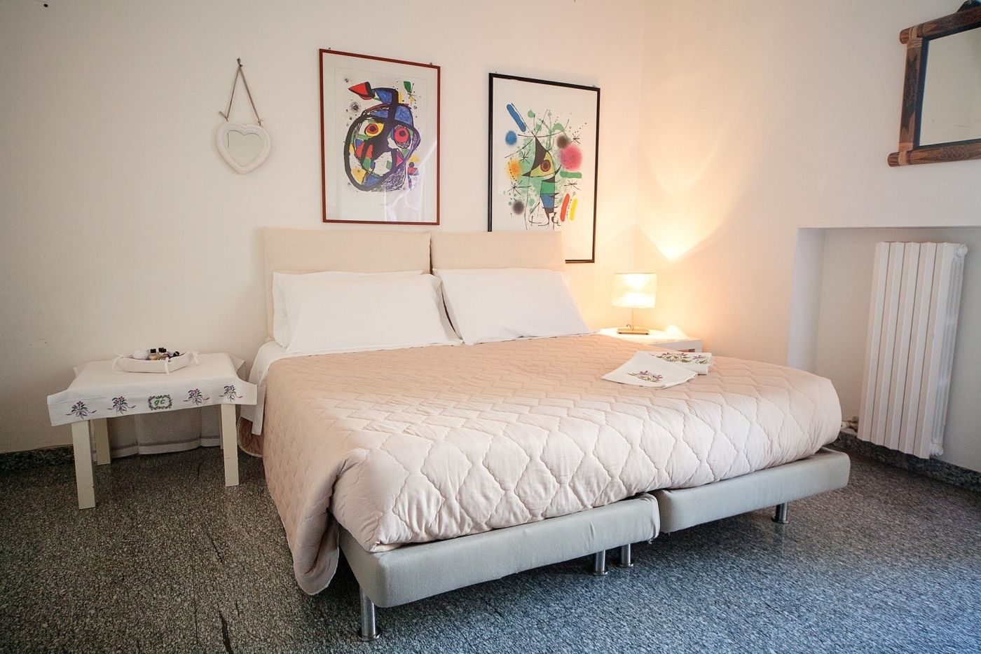 Een net opgemaakt bed met een gewatteerde spreideken, een nachtkastje met een lamp, en twee ingelijste kunstwerken aan de muur.