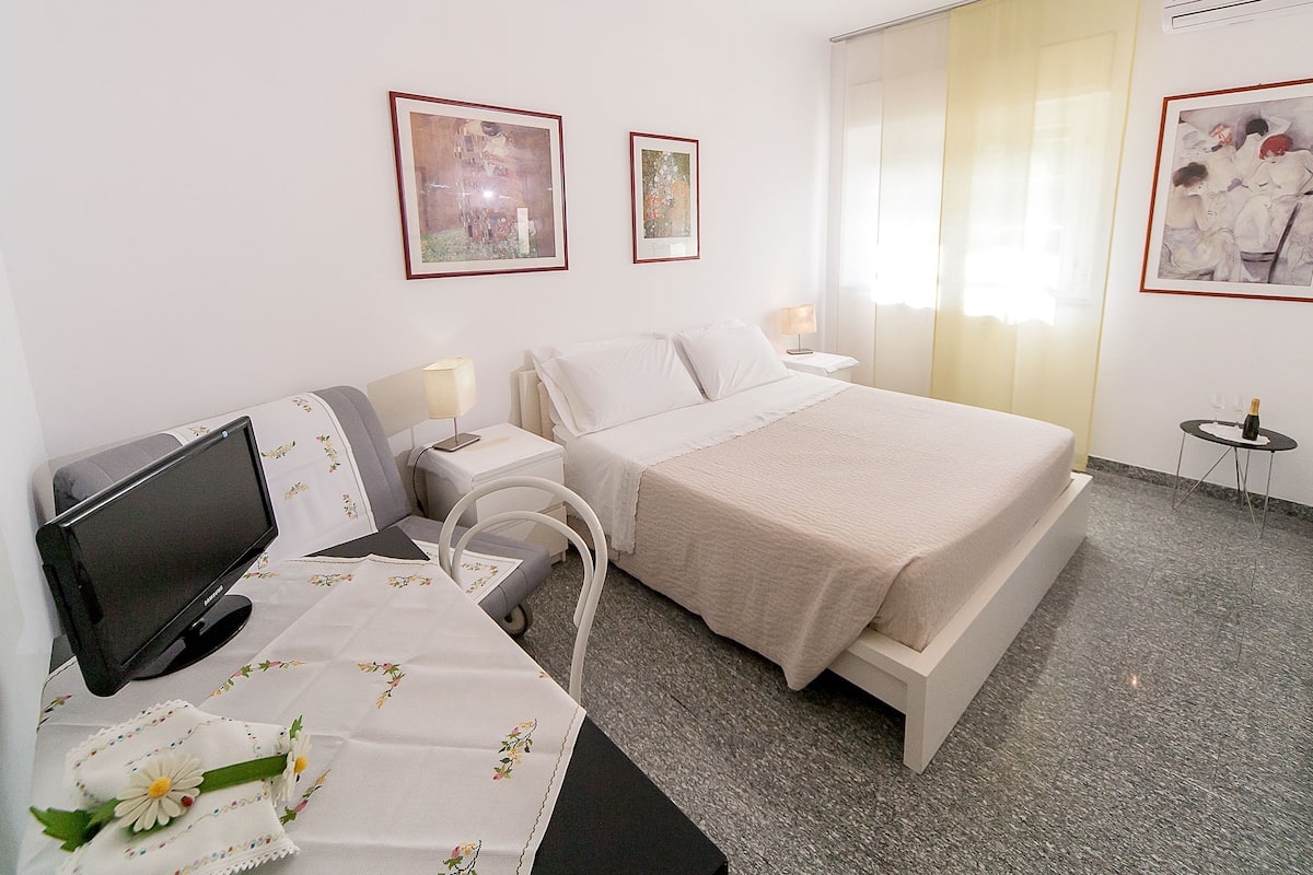 A hotel room with a bed, a desk with a computer, a chair, a small table with a lamp, and two framed artworks on the wall.
