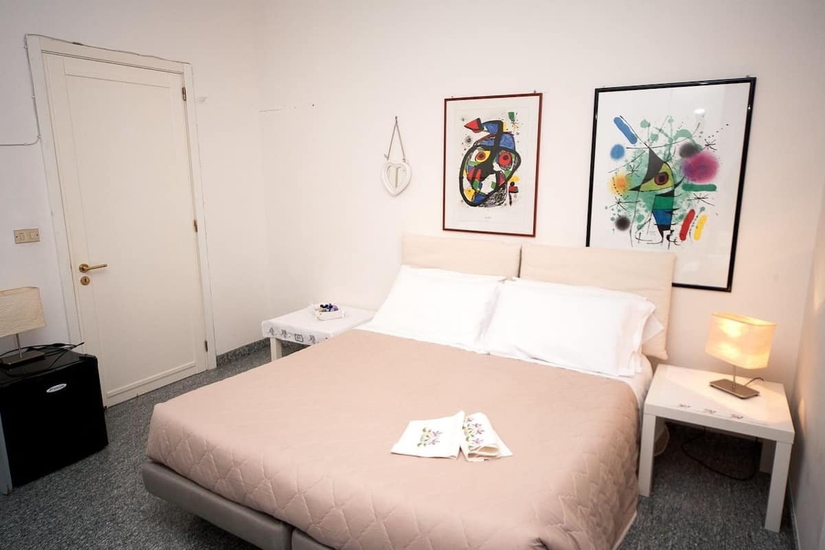 A hotel room with a bed, a bedside table, a nightstand, a lamp, a door, and two framed artworks on the wall.