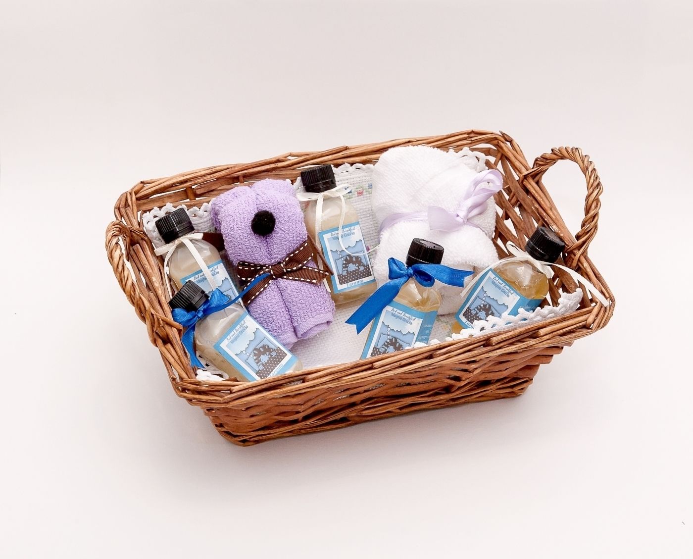 Uma cesta de vime com um brinquedo de pelúcia roxo, uma toalha branca e vários frascos de sabonete líquido com fitas azuis.