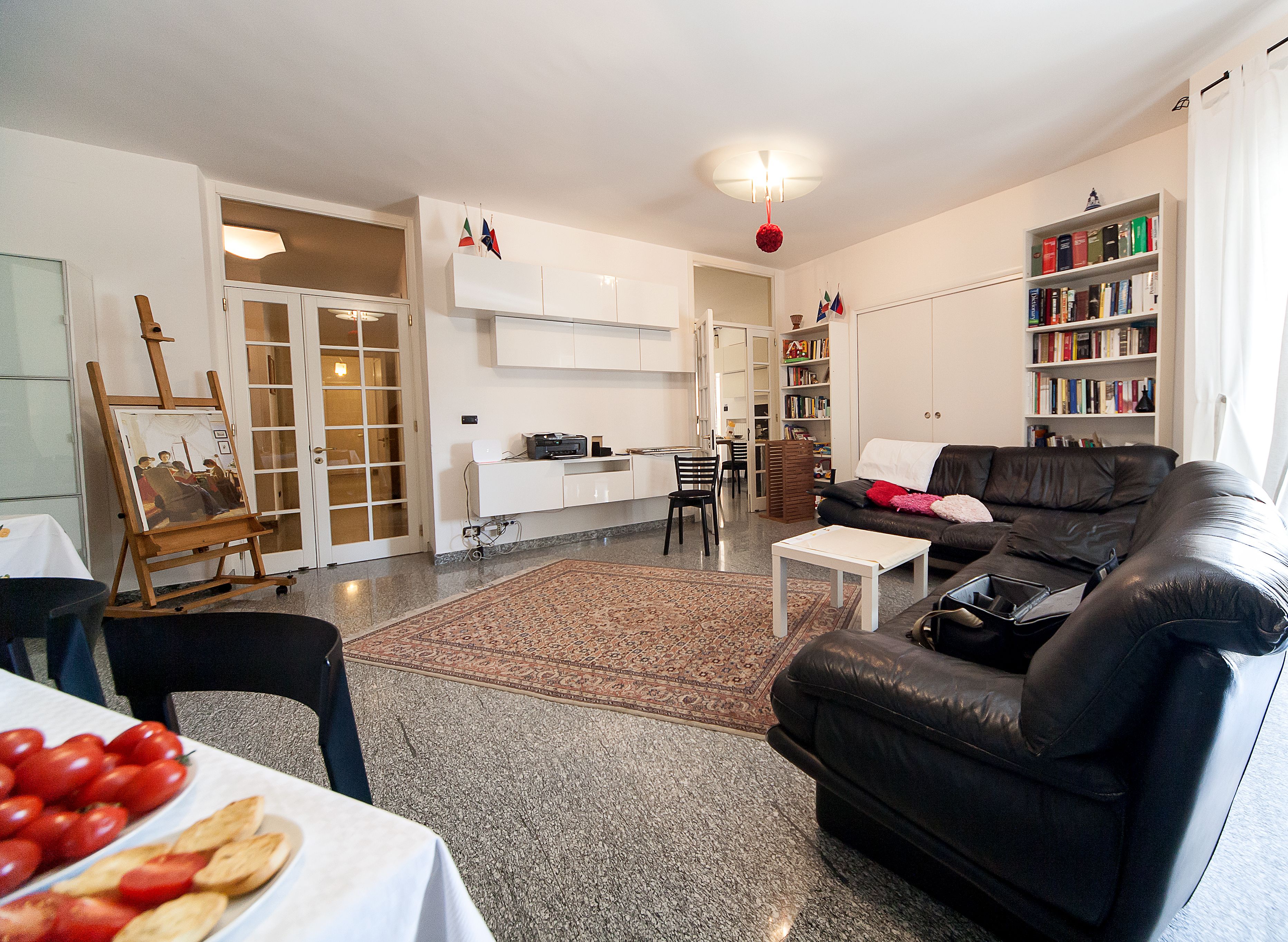 Ett vardagsrum med en svart lädersoffa, ett matbord med mat på, ett soffbord, en bokhylla och en målning på ett staffli.