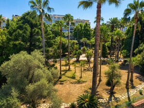A landscaped garden with palm trees and shrubs, a paved pathway, and a building in the background. - The Originals Résidence, Les Strélitzias, Antibes Juan-Les-Pins (Antibes)