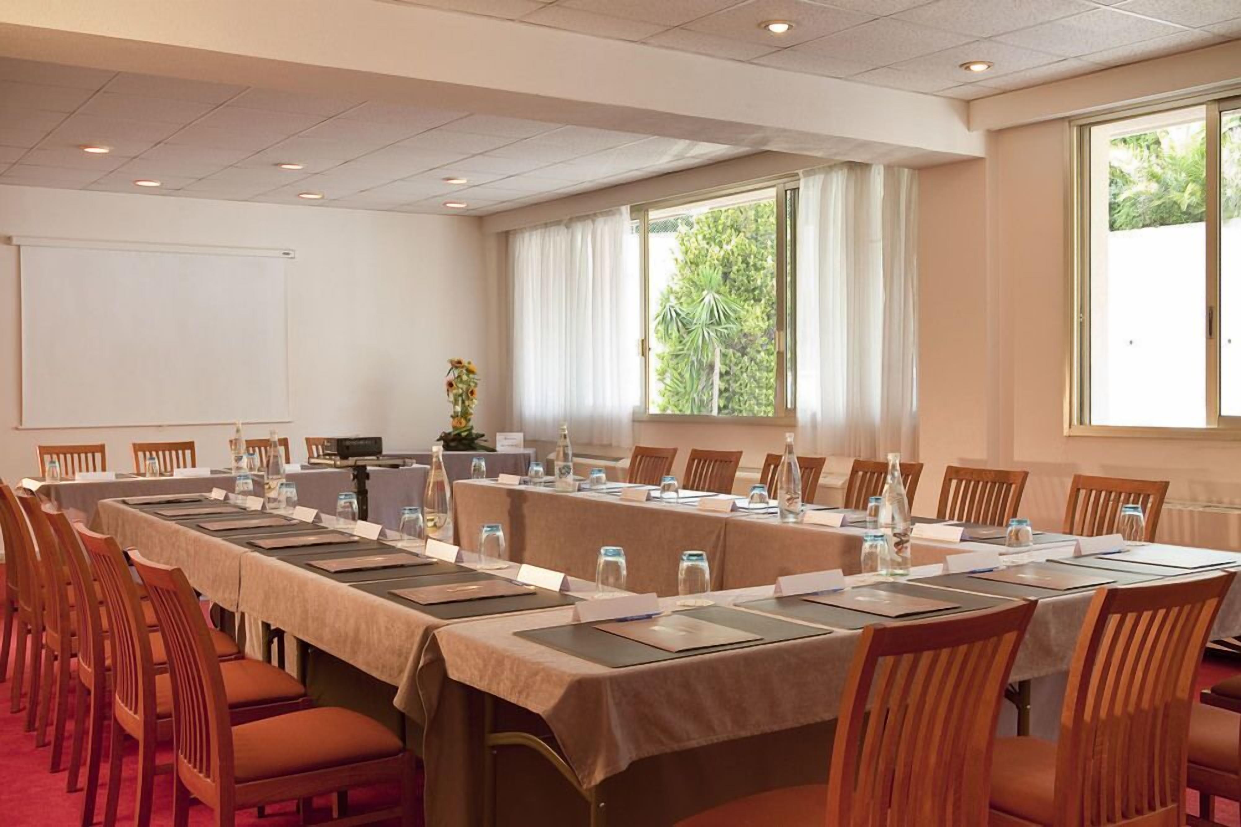 Uma sala de conferências com uma mesa comprida, cadeiras, uma tela de projetor e janelas com cortinas.