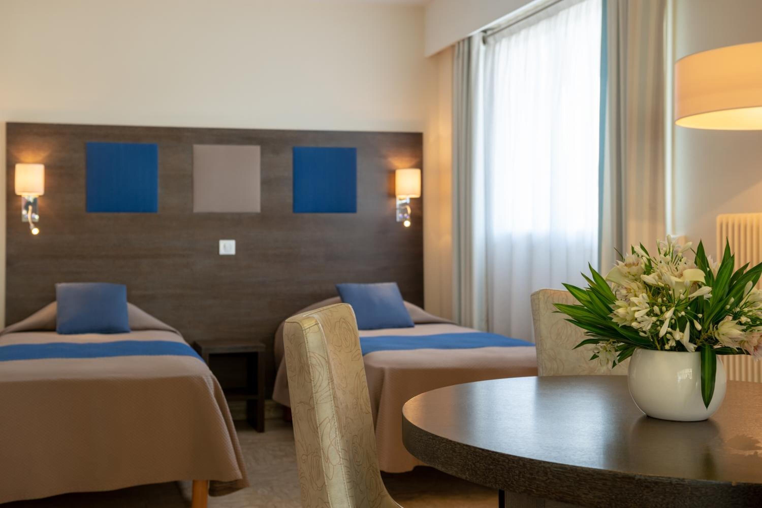 A hotel room with two beds, a small table, a vase with flowers, and a window with curtains.