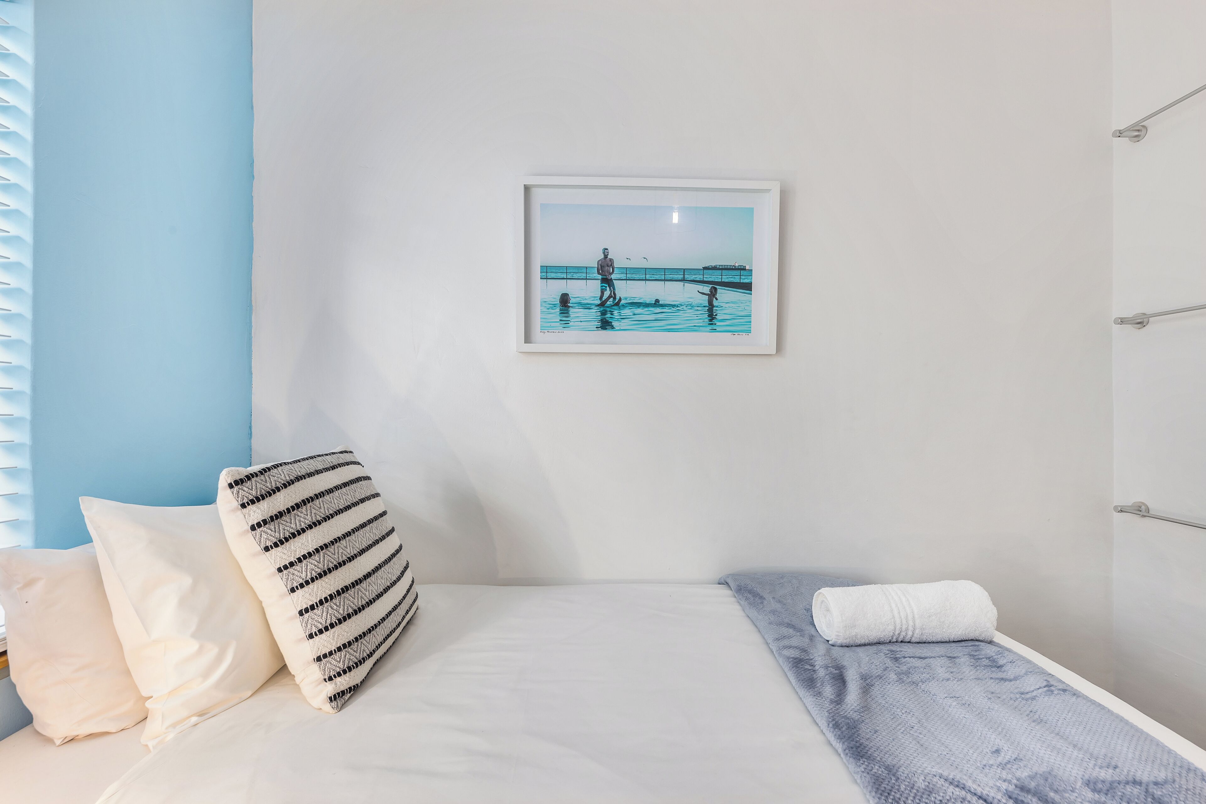 A bedroom with a bed, pillows, a framed picture of a beach scene, and a towel on the bed.