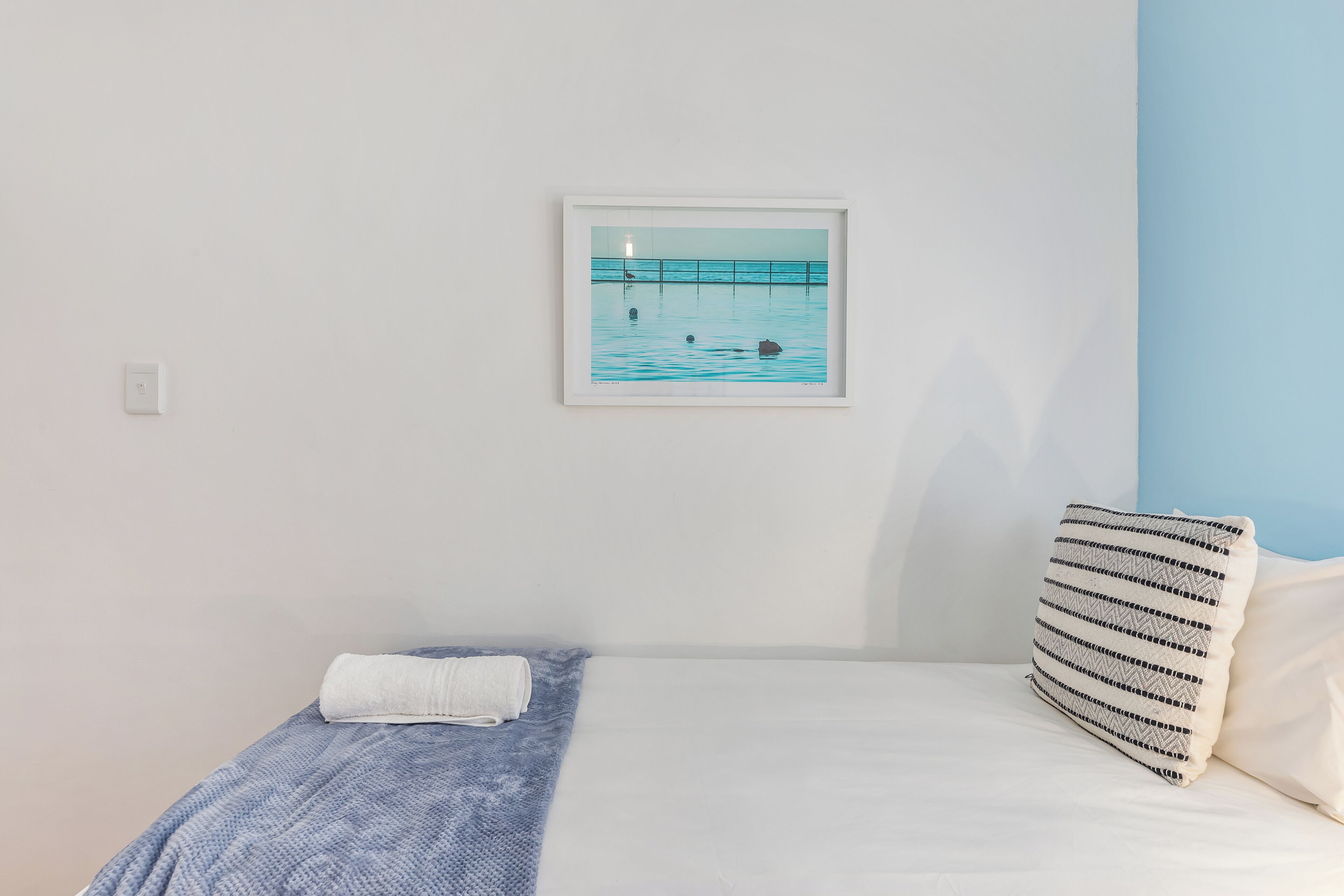 A bedroom with a bed, a pillow, a framed picture of a pier, and a wall socket.