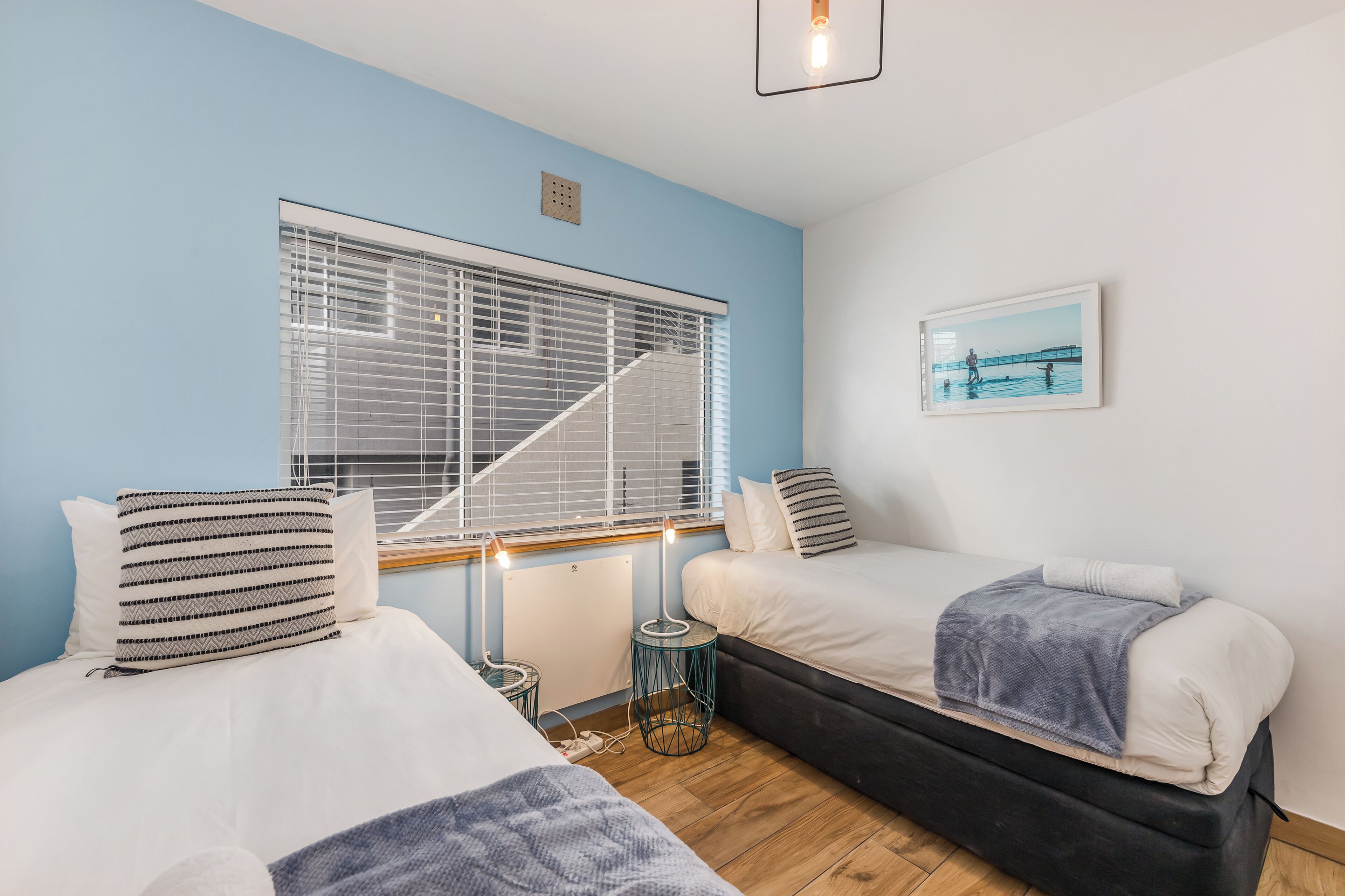 Une chambre avec deux lits, une fenêtre avec des stores, un radiateur, une petite table et un tableau au mur.