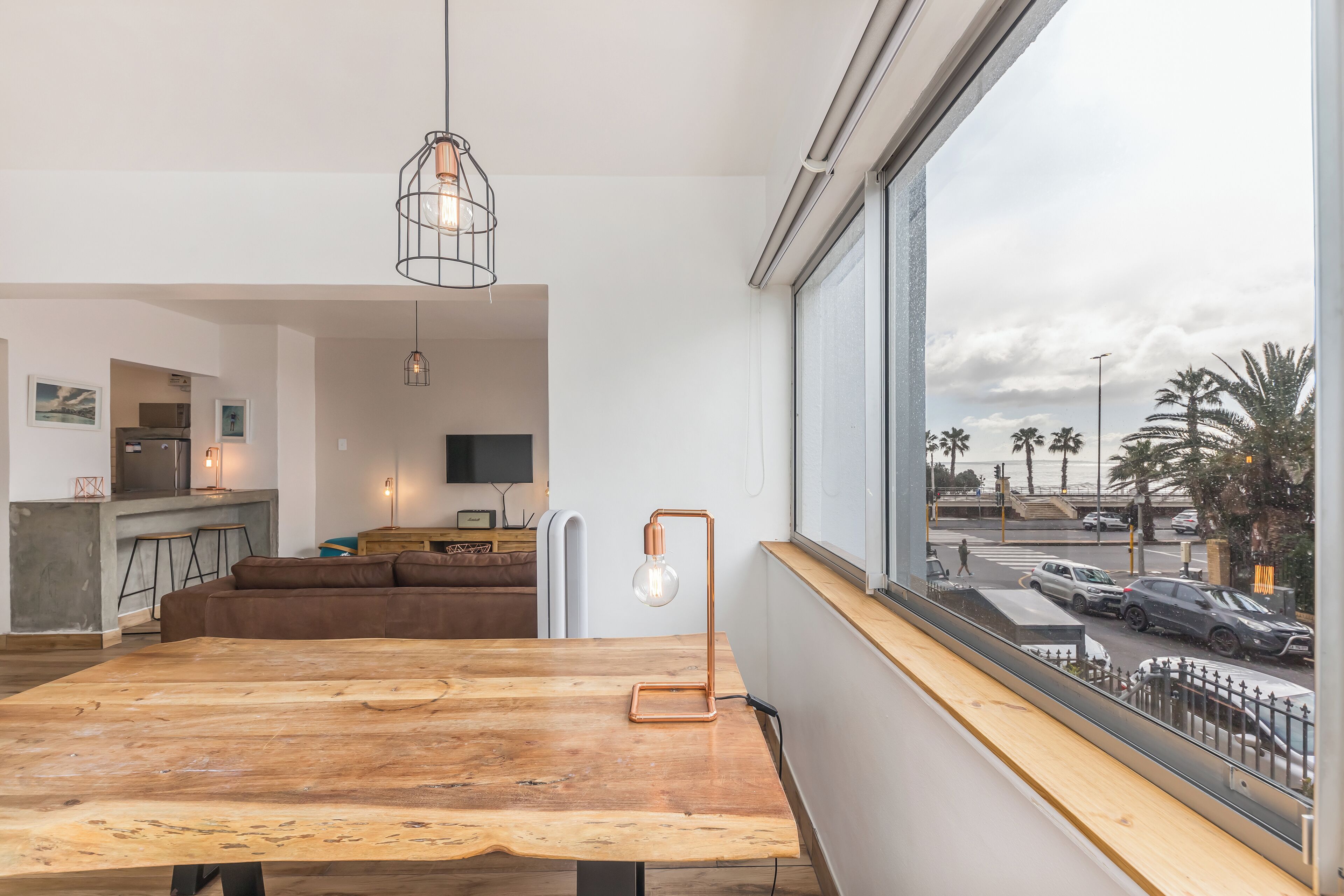A modern kitchen with a dining area, a large wooden table, and a view of the street through a large window.