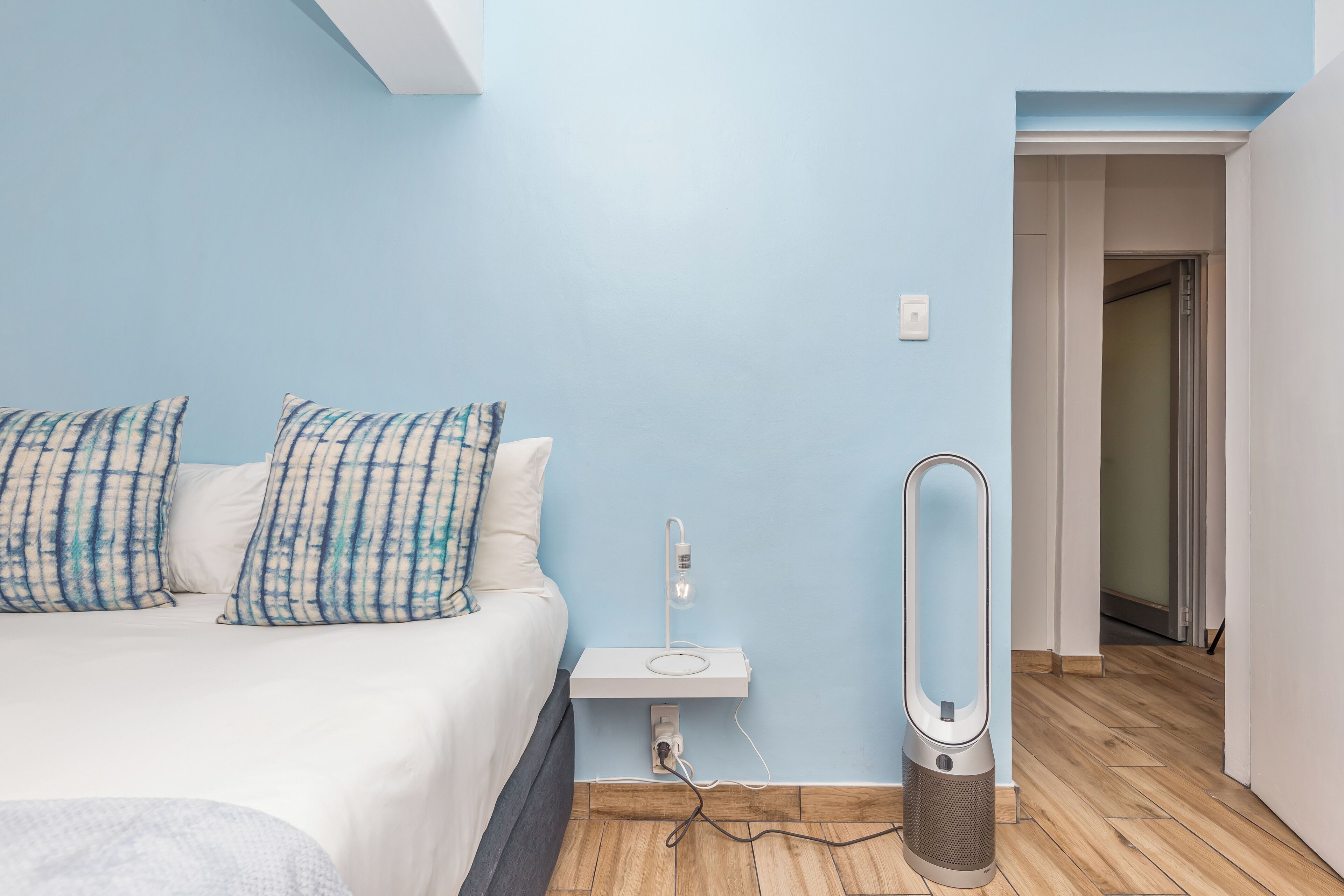 A bedroom with a bed, a bedside table, a vacuum cleaner, and a door leading to another room.