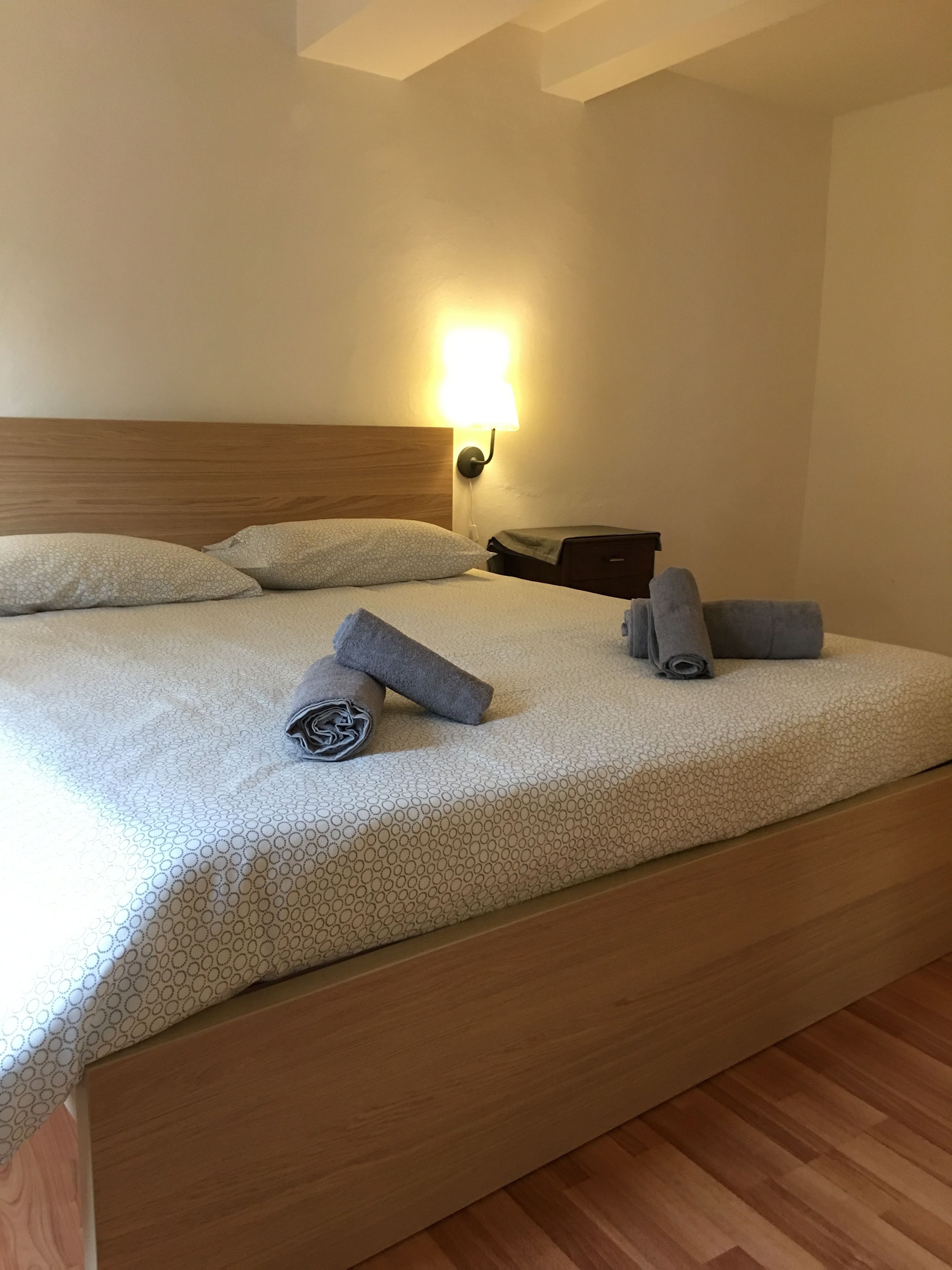 A neatly made bed with a headboard, pillows, and rolled towels. A wall-mounted lamp provides light.