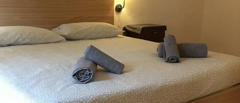 A neatly made bed with a headboard, pillows, and rolled towels. A wall-mounted lamp provides light.