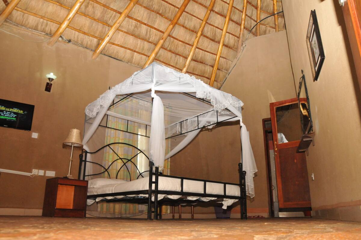 A room with a bed under a thatched roof, a nightstand with a lamp, and a wooden door.