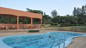 A swimming pool with a tiled edge, a covered seating area with chairs and tables, and a grassy area with trees and a hedge in the background. - Hotel Kontiki Hoima (Hoima)