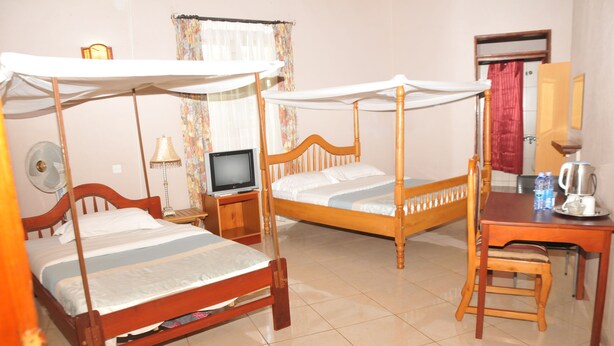 A room with two wooden beds with canopy beds, a desk with a chair, a television, a fan, and a coffee maker. - Hotel Kontiki Hoima (Hoima)
