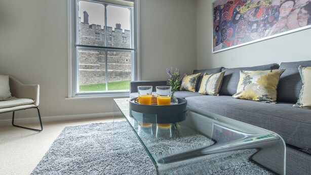 Una sala moderna con un sofá gris, una mesa de centro de vidrio con jugo de naranja y un ventanal con vista a edificios de piedra.