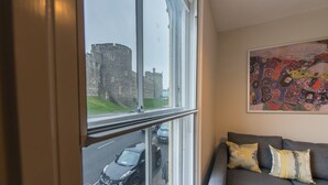 Una habitación con un amplio ventanal que da a un castillo, un sofá gris con cojines y un cuadro abstracto de colores en la pared.