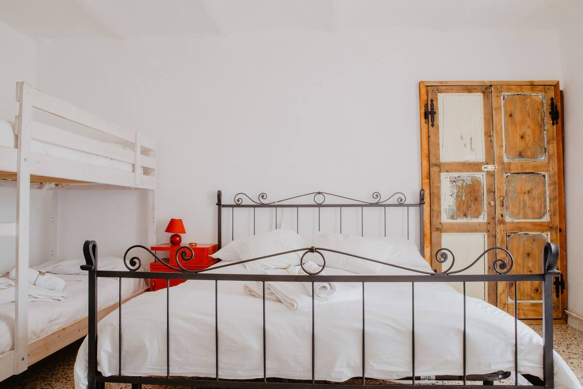A single bed with a metal frame and white bedding, a red lamp, and a wooden door with peeling paint.