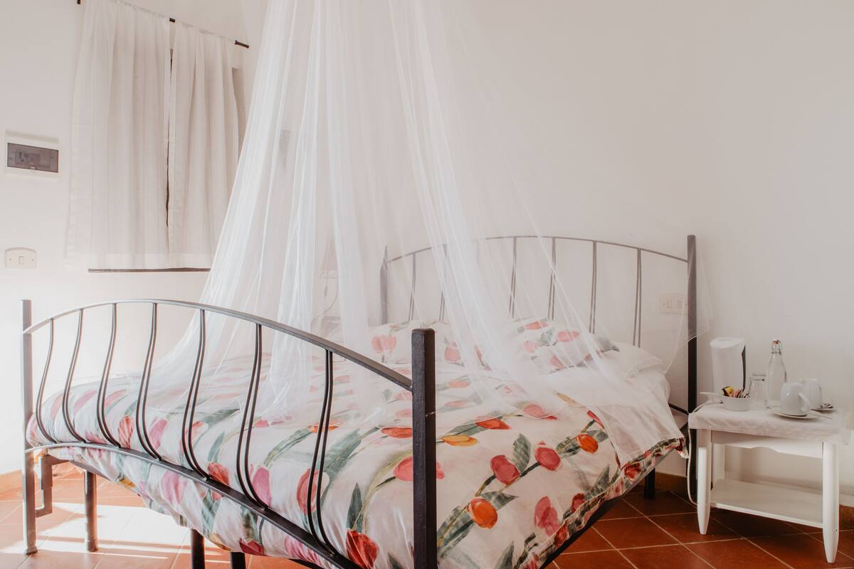 A bed with a floral bedspread, a white bedside table with a lamp, and a white curtain.