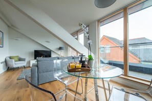 A modern living room with a glass dining table, grey sofa, and a large window offering a view of the city. - Hampden Apartments - The Elizabeth (Windsor)
