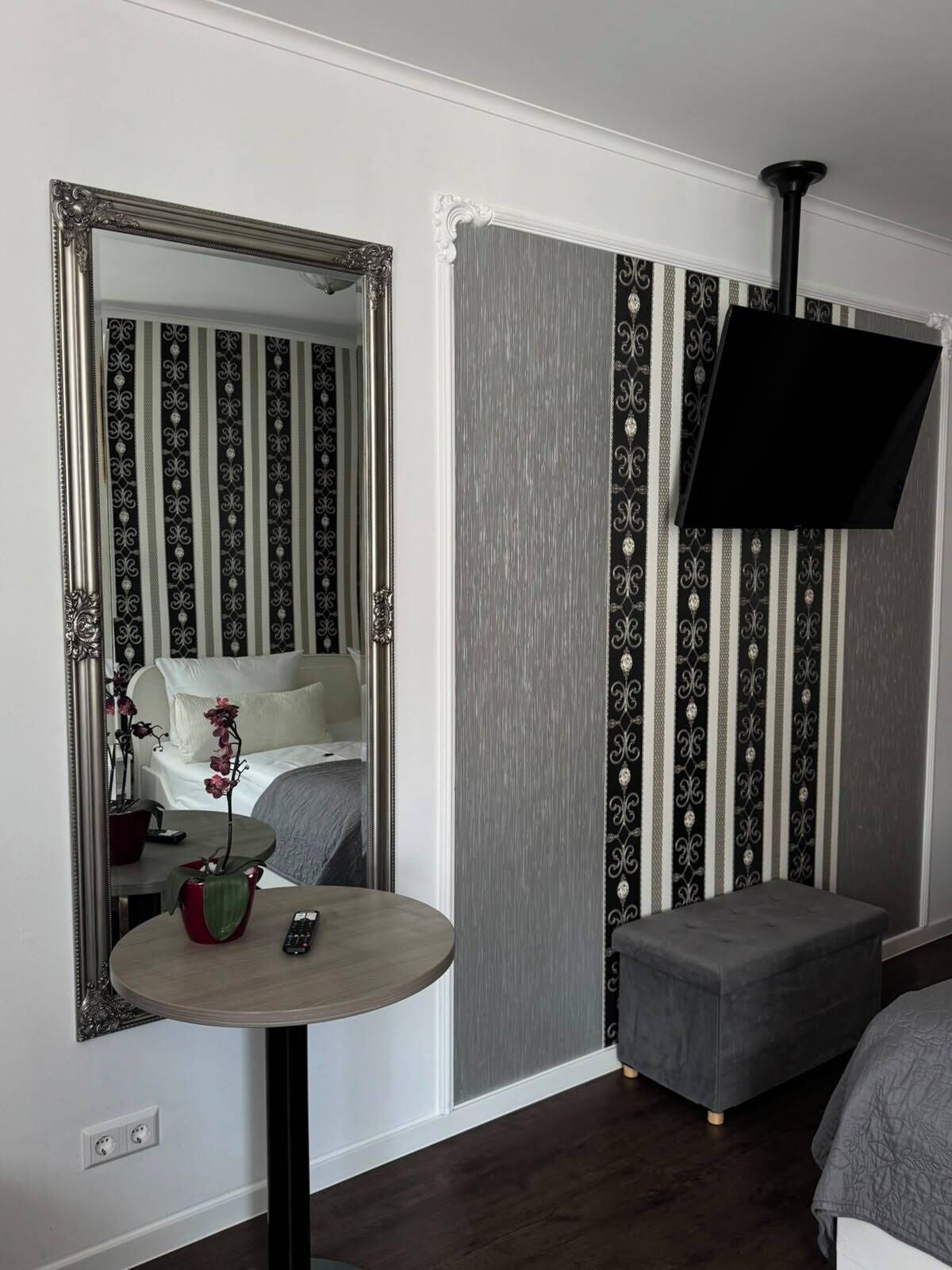 Habitación de hotel con un espejo grande, una mesita redonda con un florero y un control remoto, un otoman gris y una cama con cabecera blanca.