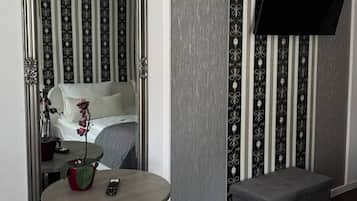 Habitación de hotel con un espejo grande, una mesita redonda con un florero y un control remoto, un otoman gris y una cama con cabecera blanca.
