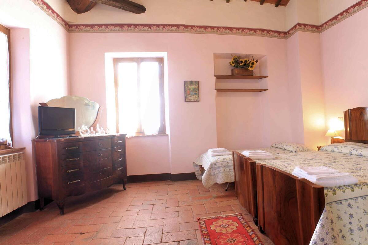 A bedroom with a wooden dresser, a television, a bed with floral bedding, a small table, and a window with sunlight.