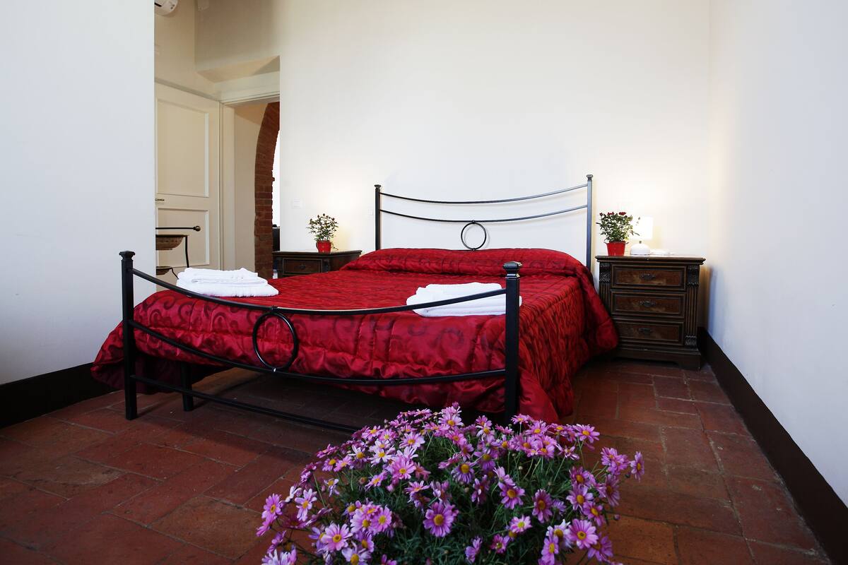 A bed with a red bedspread, a nightstand with a lamp, and a potted plant.