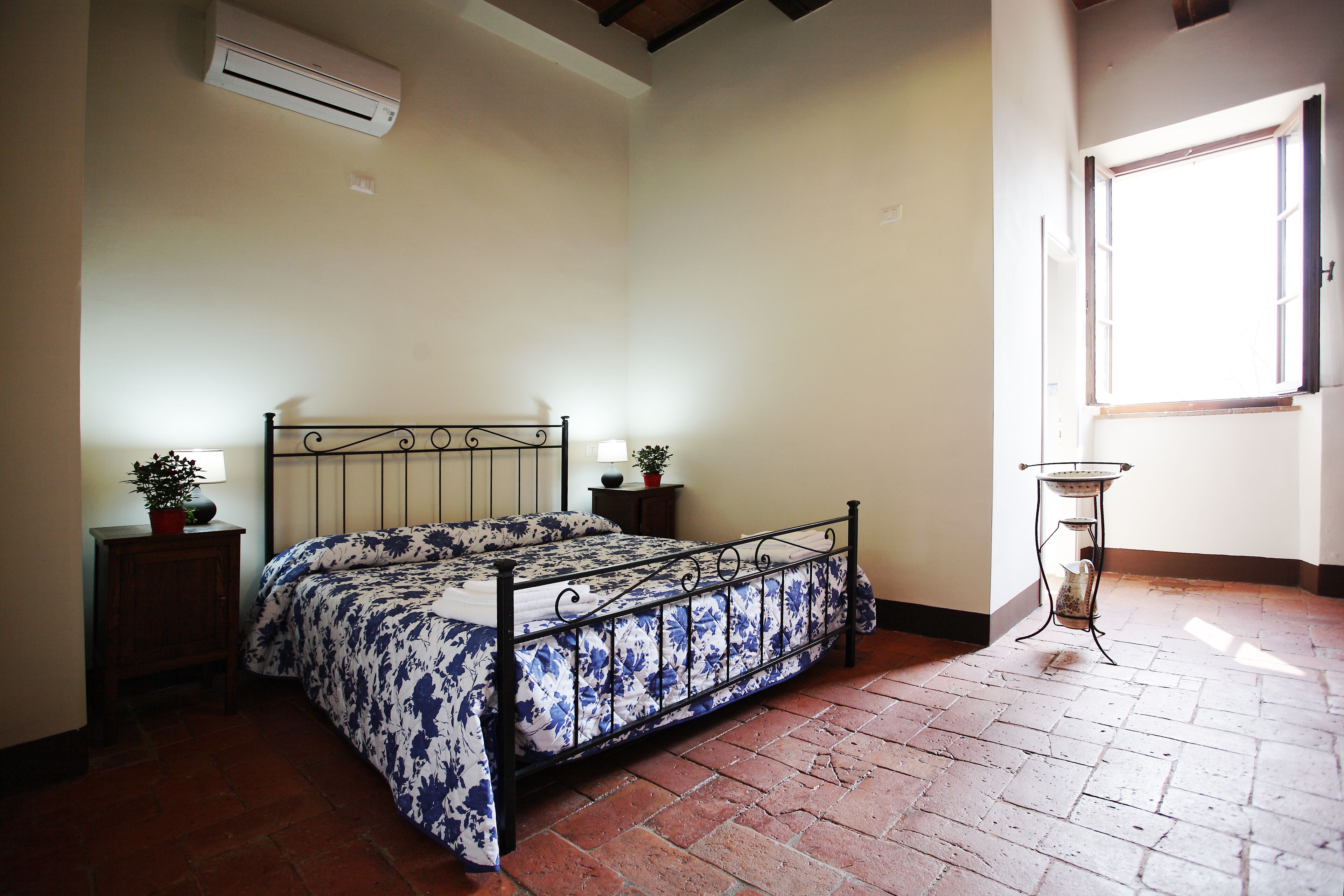 A bedroom with a metal bed frame, a brick floor, a small bedside table with a lamp, and a window with a view of the outside.
