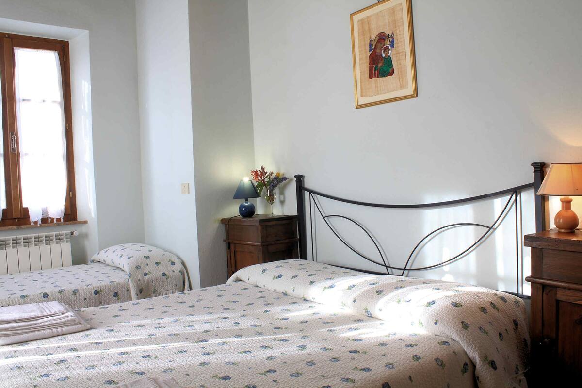 A bedroom with two beds, a window with curtains, a bedside table with a lamp, and a framed picture on the wall.