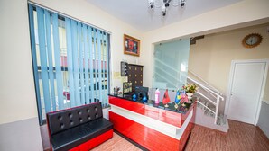 A reception area with a red and white desk, a black sofa, a blue curtain, a framed picture on the wall, and a staircase leading to an upper floor. - Hotel Primavera (Sarandë)