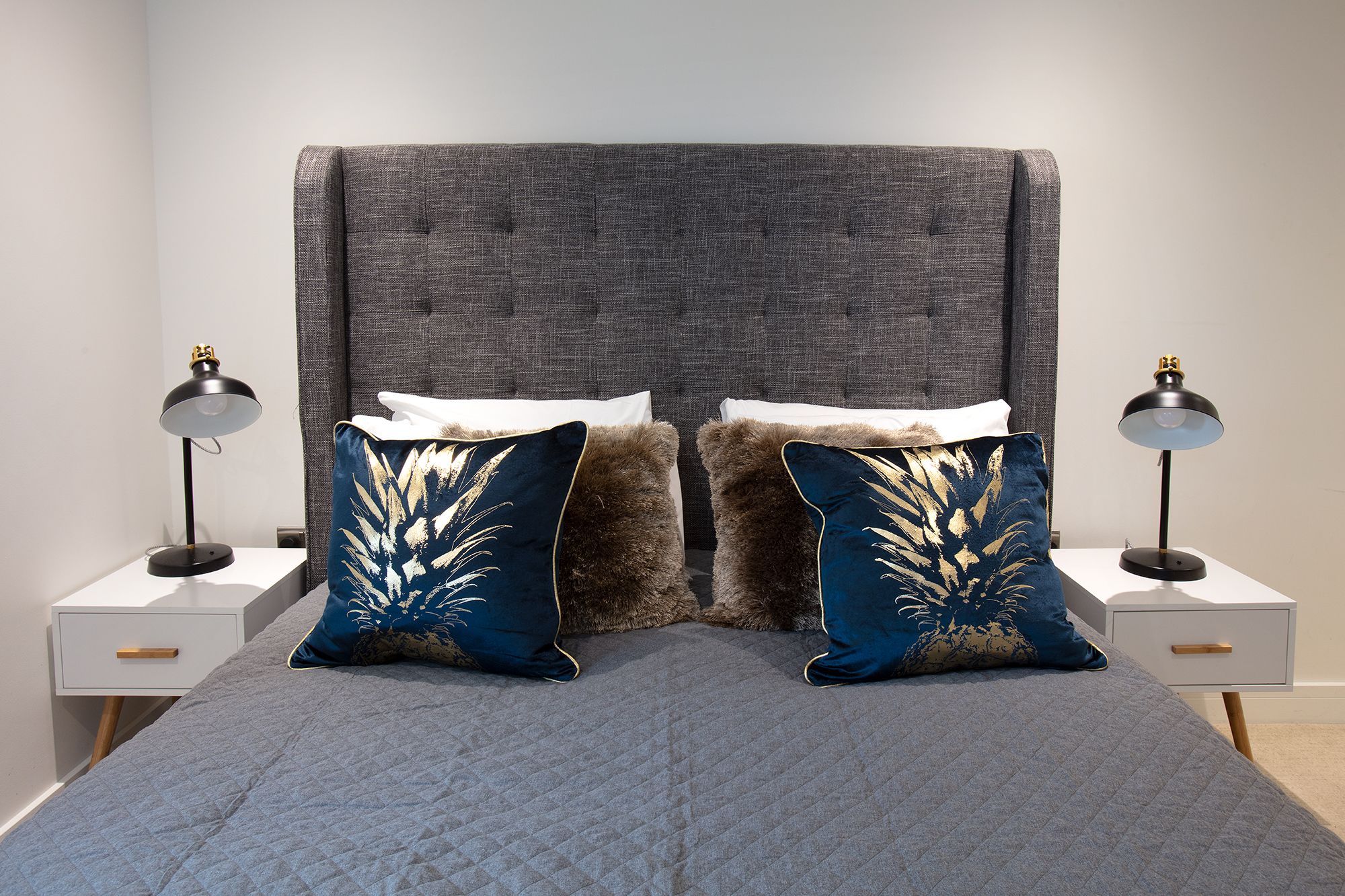 A bed with a grey quilted cover, two matching pillows with a gold leaf design, and a grey upholstered headboard.
