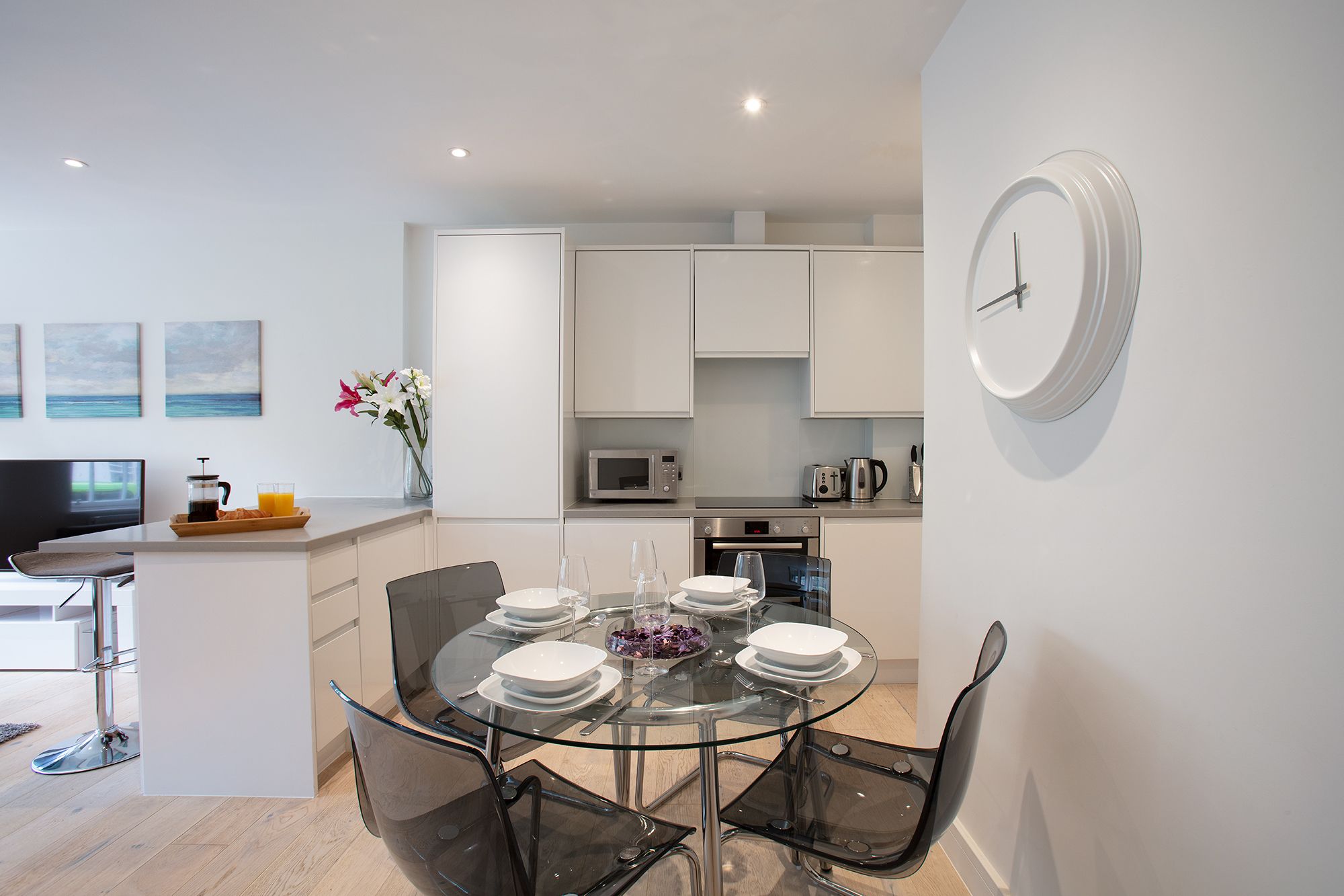 A modern kitchen with white cabinets, a microwave, and a coffee maker. There is a dining area with a glass table and chairs.
