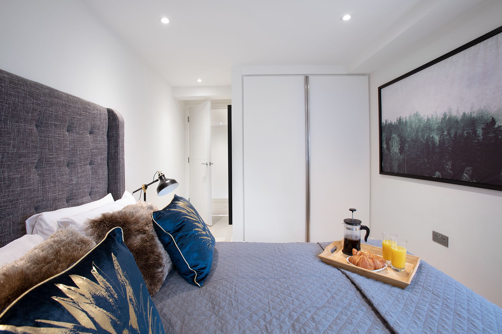 A modern bedroom with a large bed, a coffee table with breakfast items, and a forest-themed painting on the wall.