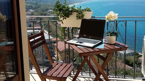 Balcón con una computadora portátil, sillas de madera y plantas en macetas, con vistas a la costa.