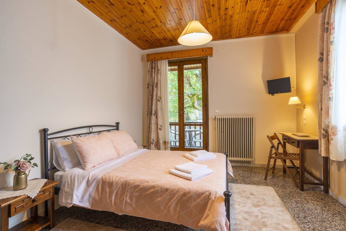A bedroom with a wooden ceiling, a bed with pink bedding, a bedside table with a vase, a desk with a chair, and a TV on the wall.