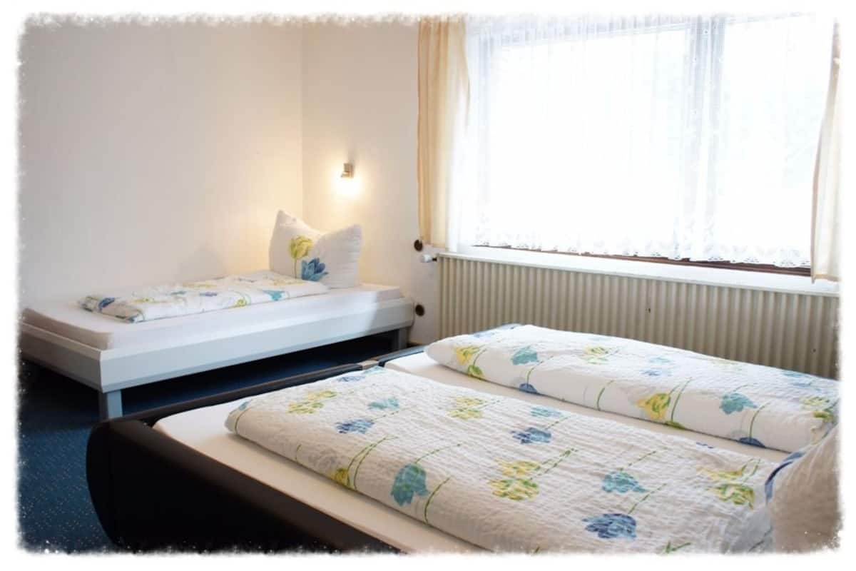 A small, single-bed room with a window, a radiator, and a bedside table with a lamp.