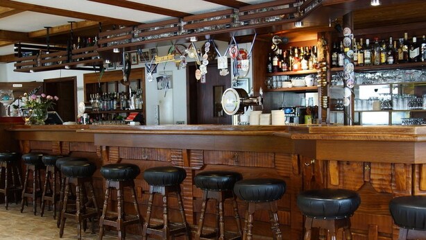 A bar area with wooden stools, a bar counter, and shelves stocked with bottles and glasses. - Höfener Wirtshaus (Monschau)